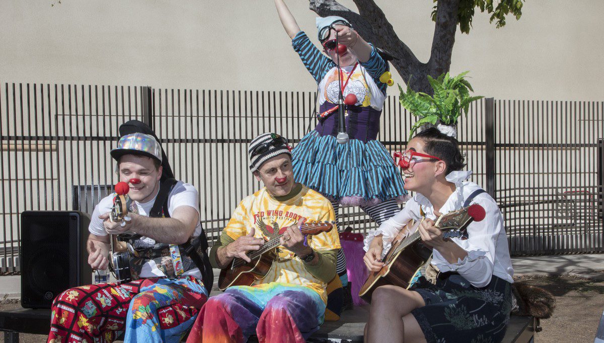 Four clown musicians play guitars and ukuleles outdoors while a fifth performs behind them.